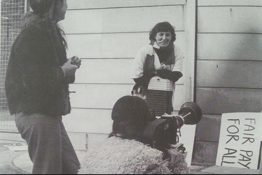 black and white photograph showing a woman being filmed by a camera with a sign reading 'fair pay for all'