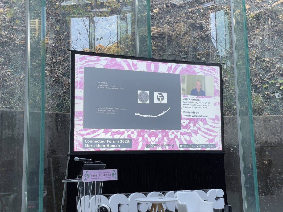 photograph of a television screen into a glass building. On the screen is Prof. Neal White giving an online presentation