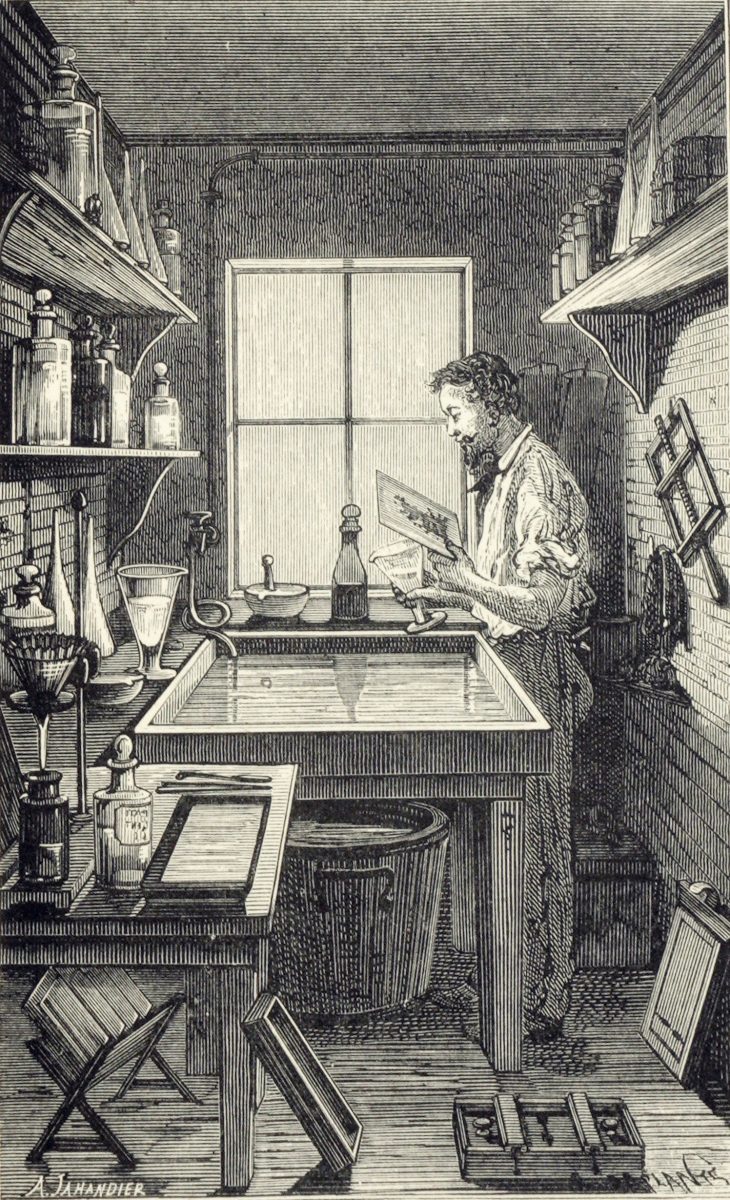 Etching or engraving showing a man in a photographic darkroom space. He is looking at a fresh photographic print which he has presumably just taken out of the developer.