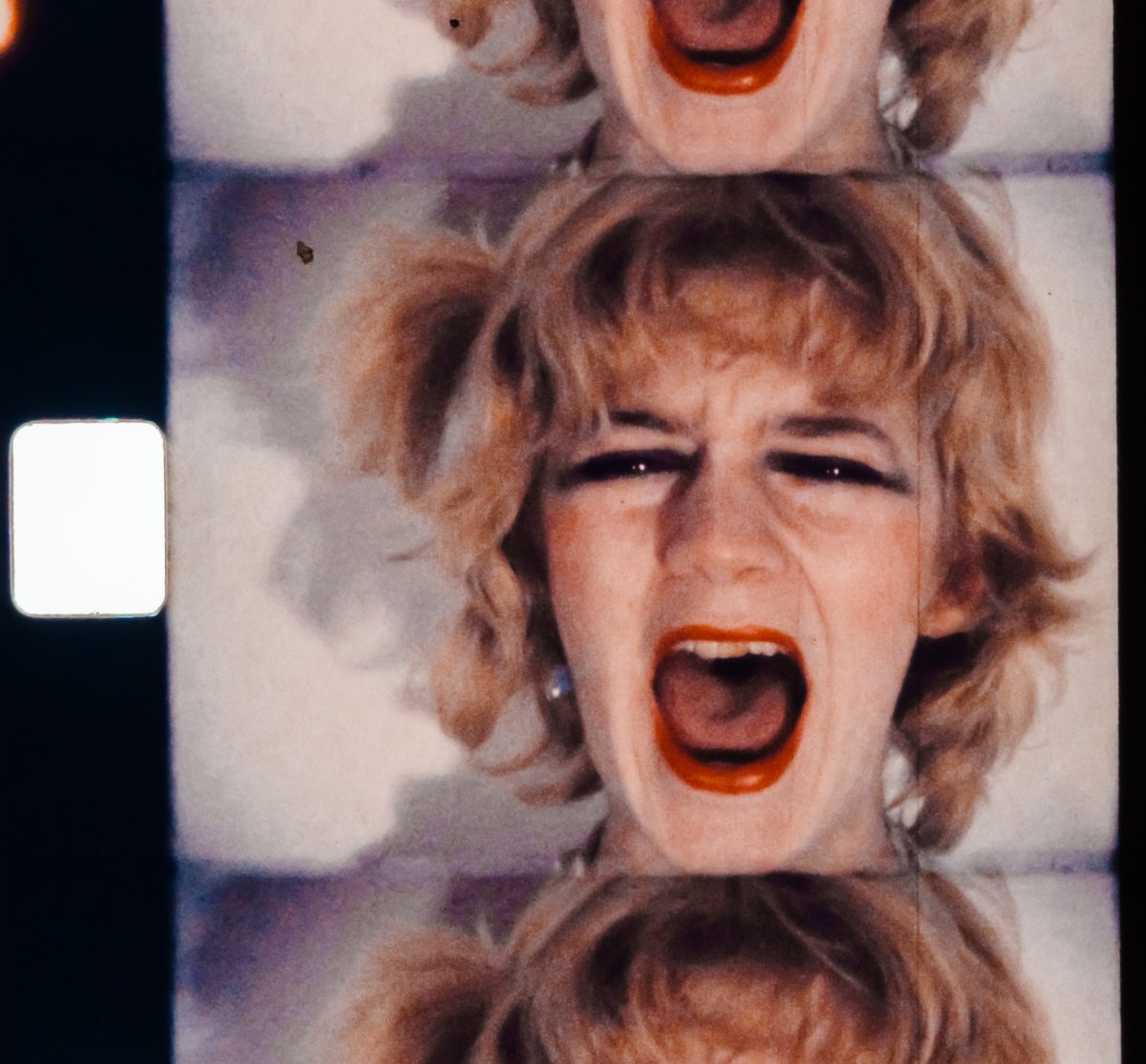 film still showing a woman with blonde hair and red lipstick, screaming. The image shows that it is taken from analogue film. 