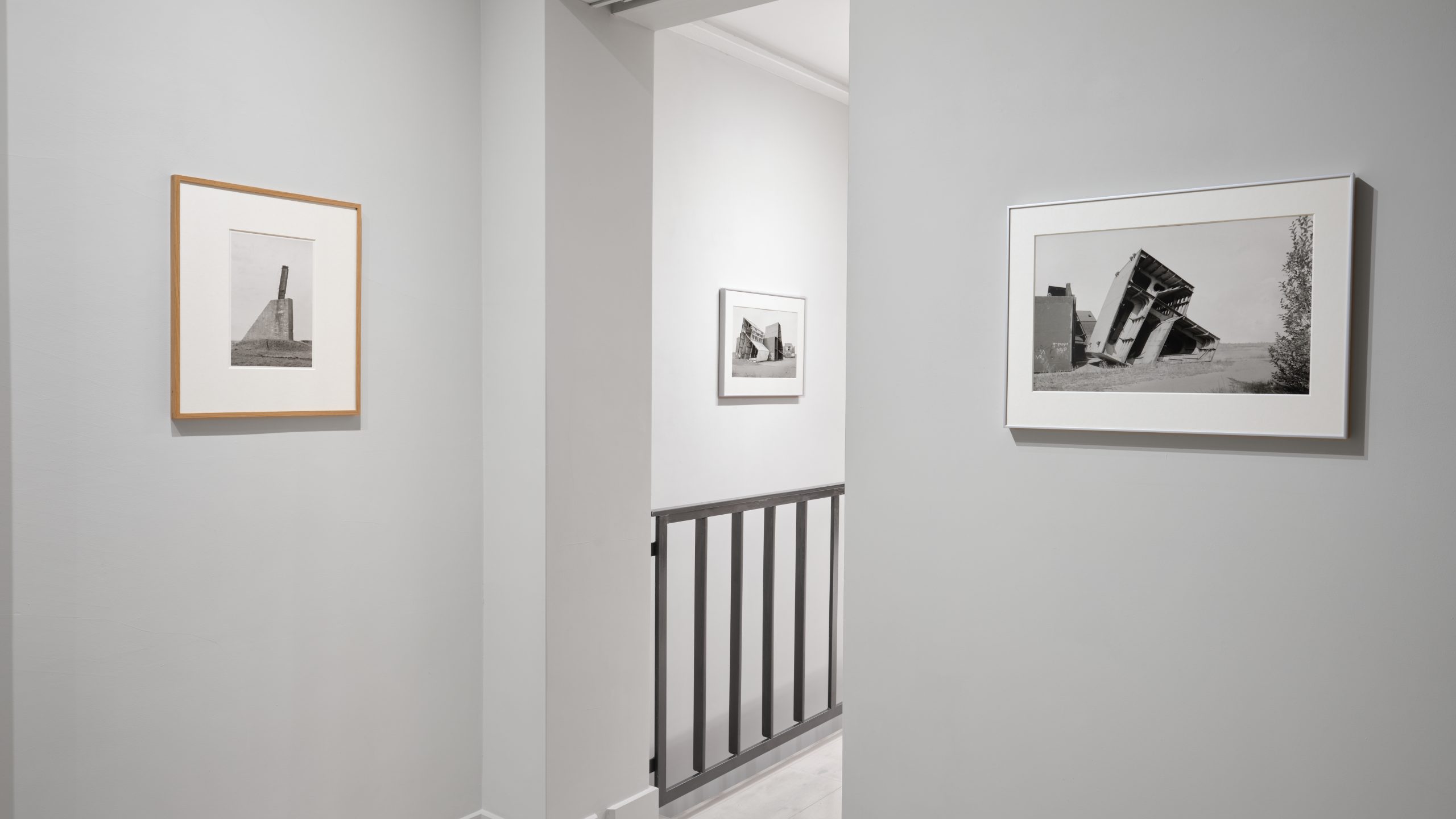 Installation view of a gallery space. Shows three framed photographs. To the left, a photograph of an architectural tower. To the right, a photograph of an anonymous building or sculpture. In the middle, a second photograph of an an anonymous building or sculpture.
