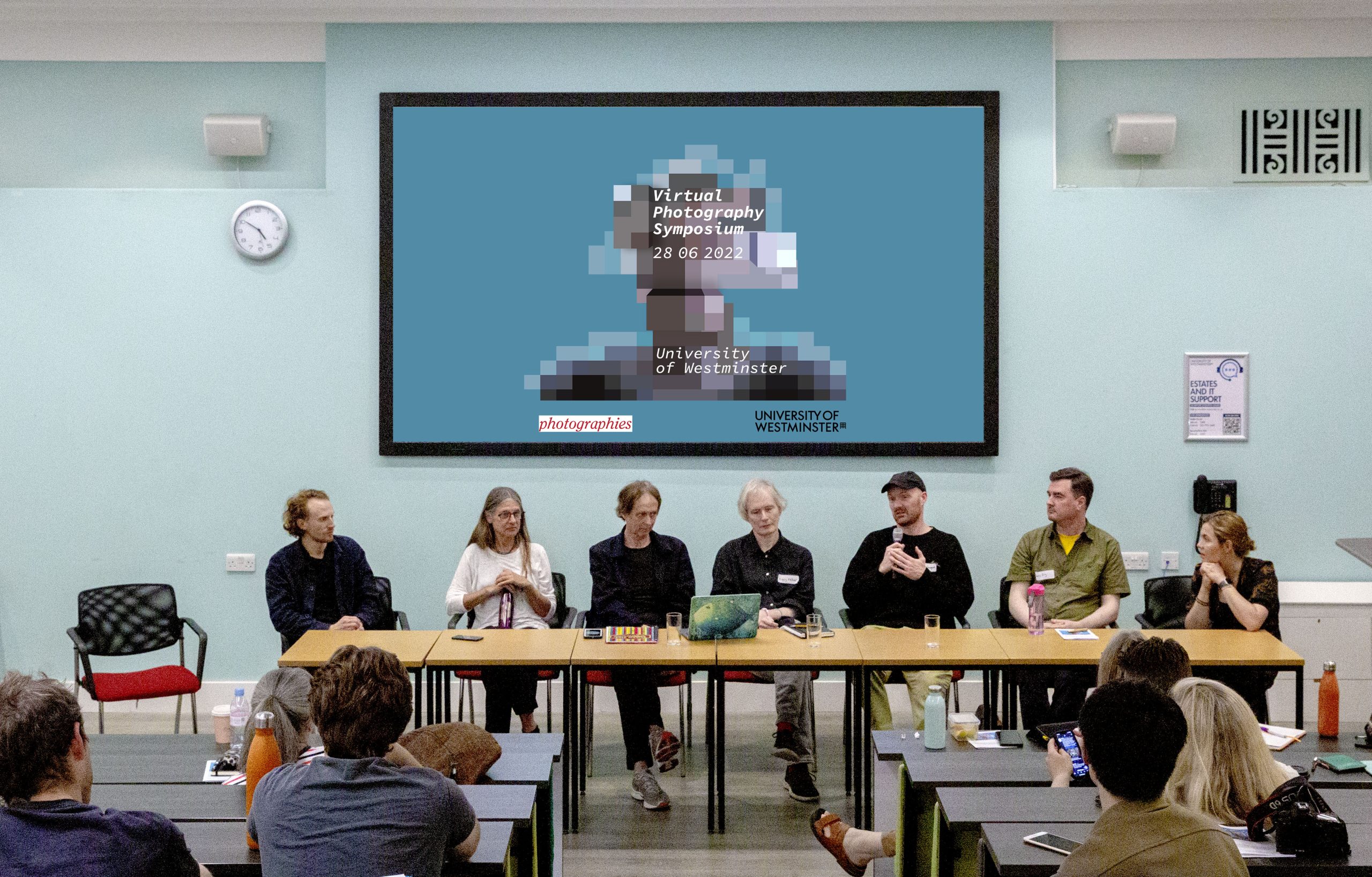 Photography of a panel discussion from the conference 'Virtual Photography Symposium'. Seven people seated at a long table, poster for the conference projected on a screen above.