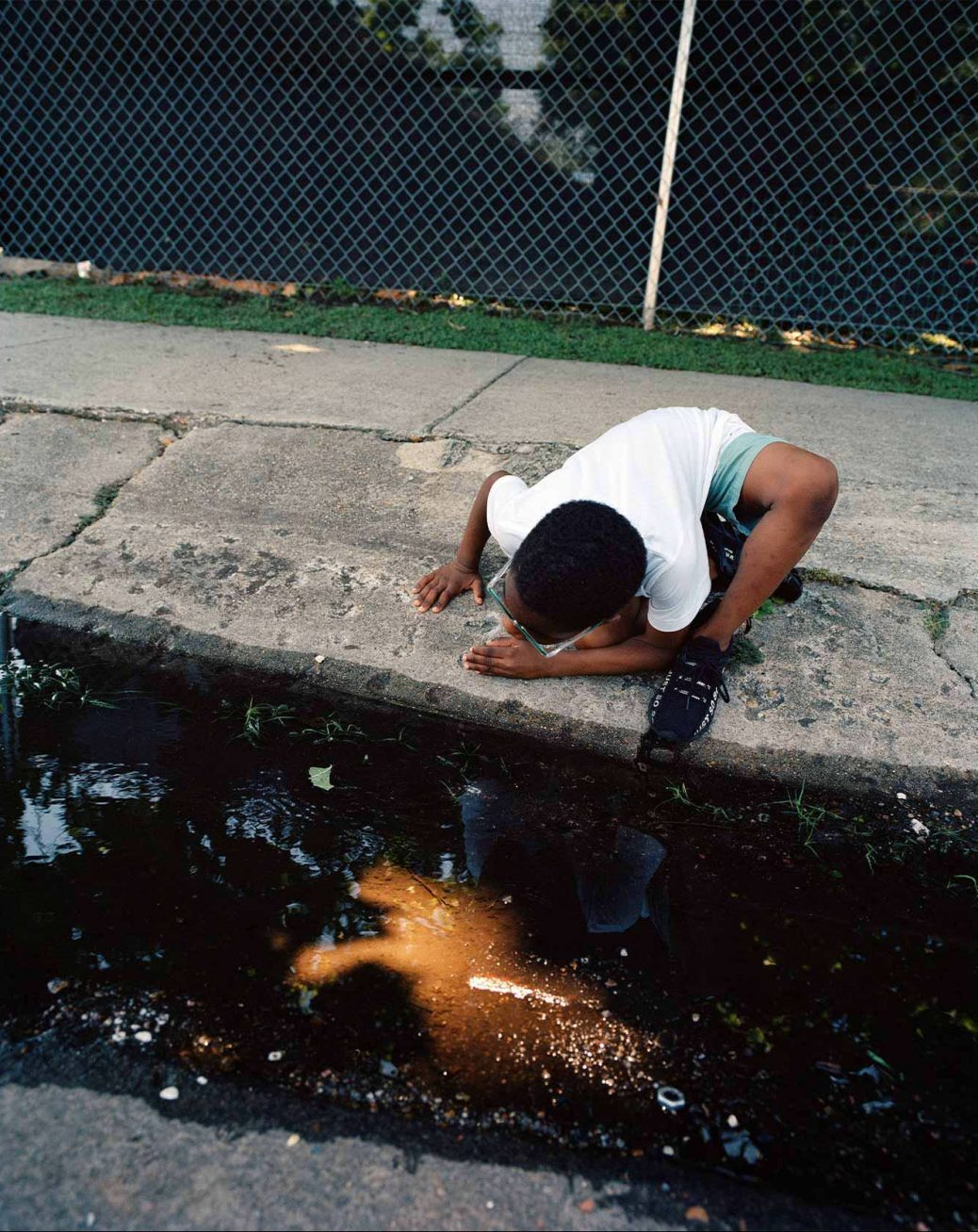 colour photograph of a boy crouched down on a pavement looking at a puddle in the street